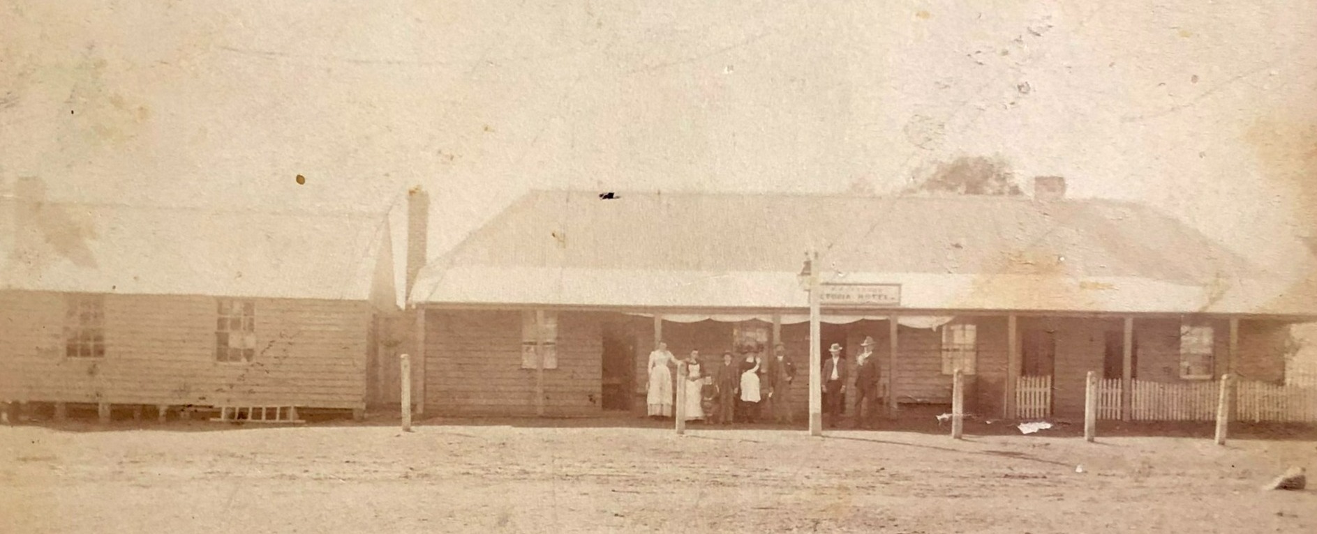 A photograph of the Victoria Hotel, now known as the Old Vic. A group of seven people are standing under the front verandah looking to the camera 